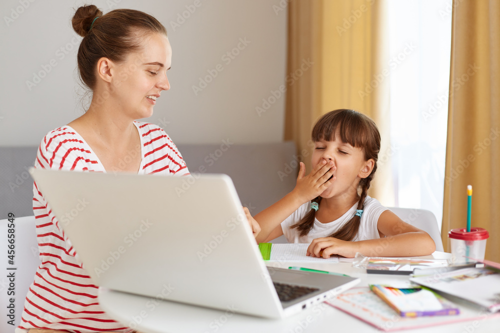 Indoor shot of sleepy tired schoolchild sitting at table in living room, mother helping her daughter with lessons, posing in front of portable computer, kid yawning. Indoor shot of sleepy tired schoolchild sitting at table in living room, mother helping her daughter with lessons, posing in front of portable computer, kid yawning.