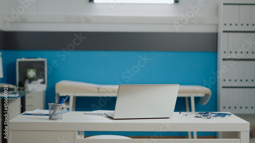 Close up of white desk with laptop and medical instruments in empty doctors office at clinic. Nobody in healthcare cabinet with stethoscope, bed for consultation and document files © DC Studio