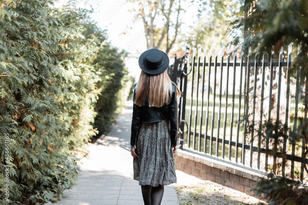 Beautiful woman with a fashion black hat in a leather jacket with a dress walks in the park on an autumn sunny day, back view