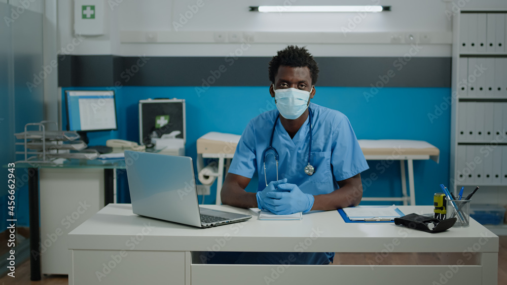 Portrait of young person working as nurse in cabinet at healthcare facility. Medical assistant sitting at white desk looking at camera with face mask, gloves, uniform and stethoscope Portrait of young person working as nurse in cabinet at healthcare facility. Medical assistant sitting at white desk looking at camera with face mask, gloves, uniform and stethoscope