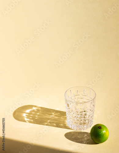 an art creative template of empty crystal glass and lime under hard sunlight, glitter reflection on beige background, long harsh rectangular and oval shadows, copy space