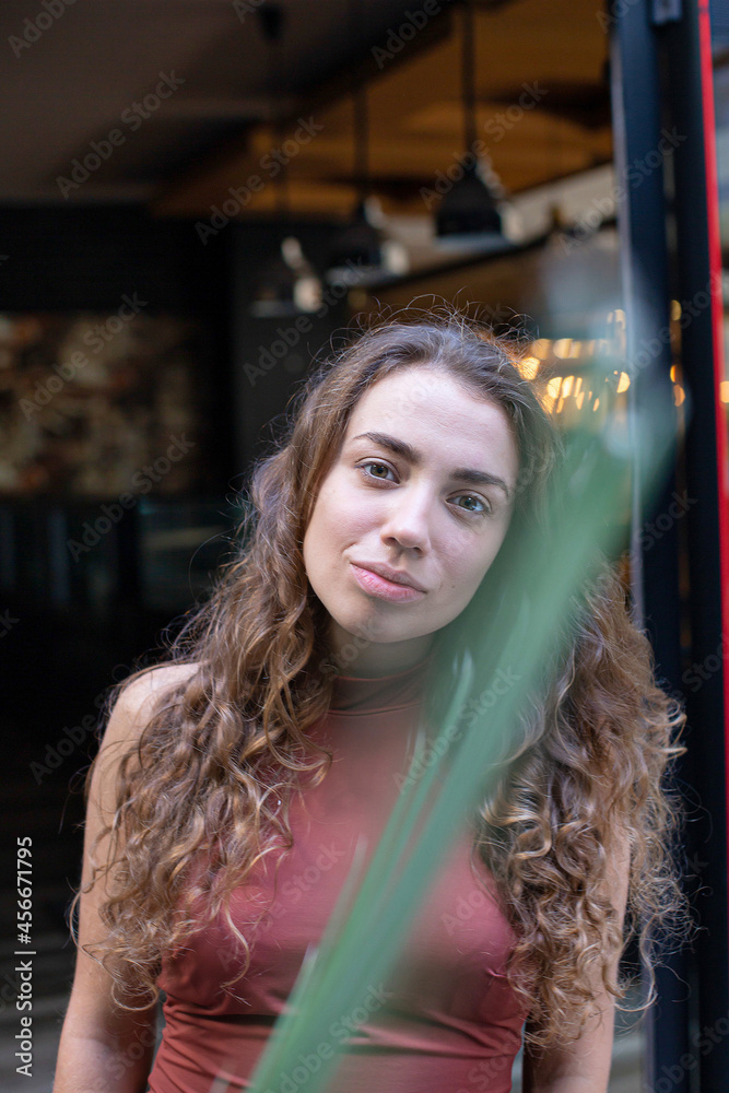 European girl with dark and curly hair with restaurant behind and palm leaf in front