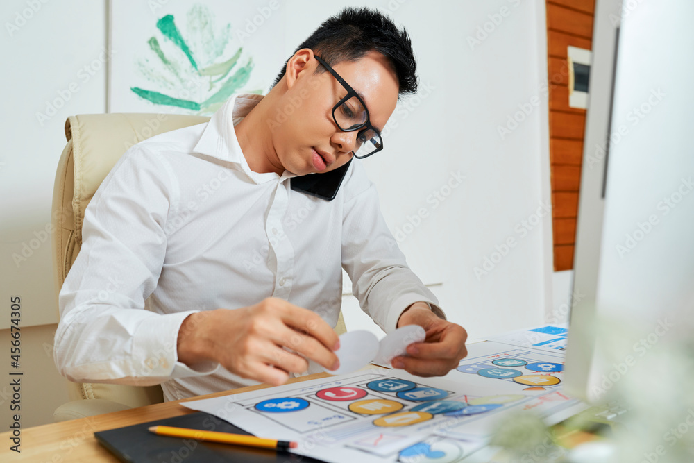 Graphic designer talking on phone with customer or colleague when working on layout of mobile application Graphic designer talking on phone with customer or colleague when working on layout of mobile application