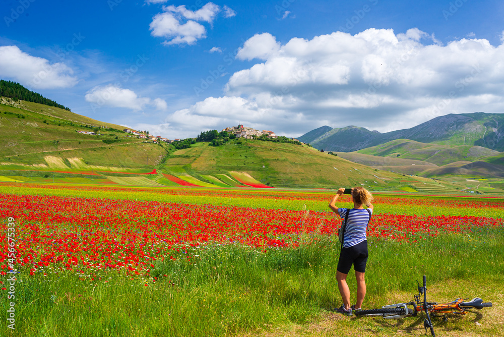 Woman cycling mtb in blooming cultivated fields of Castelluccio di Norcia highlands, famous colourful flowering plain in the Apennines, Italy. Agriculture of entil crops and red poppies.