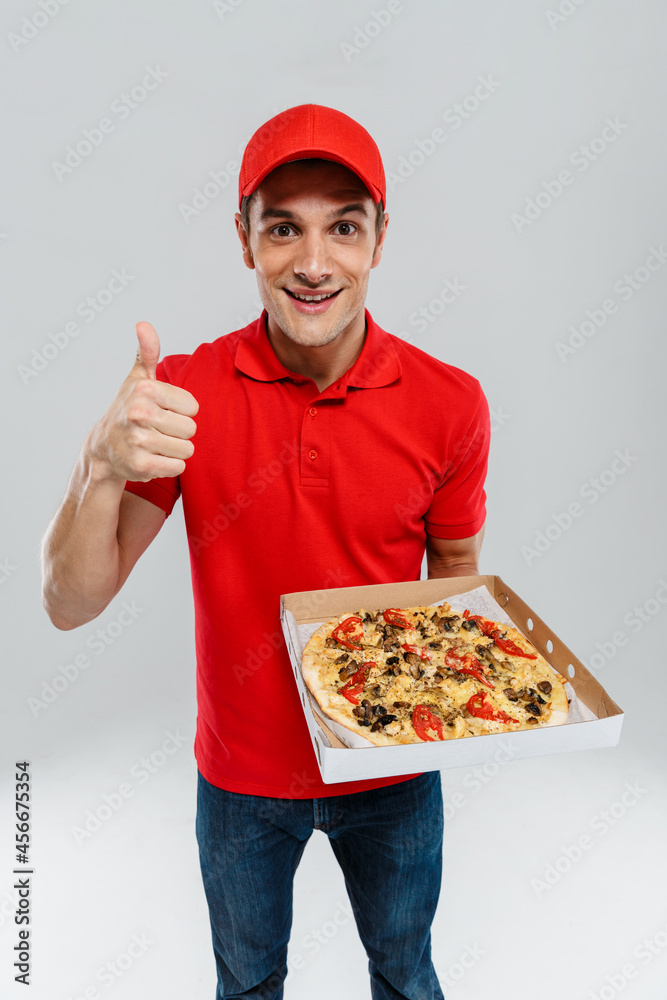 Young delivery man in uniform showing thumb up while showing pizza Young delivery man in uniform showing thumb up while showing pizza