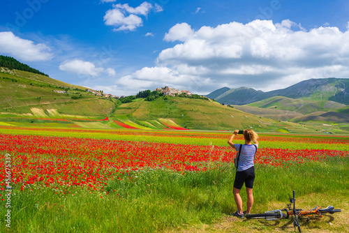 Fotografie Woman cycling mtb in blooming cultivated fields of Castelluccio di Norcia highlands, famous colourful flowering plain in the Apennines, Italy