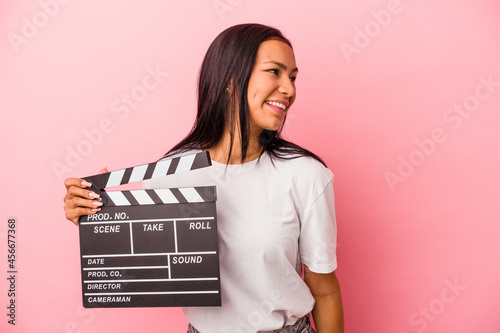 Young latin woman holding clapperboard isolated on pink background  looks aside smiling, cheerful and pleasant.