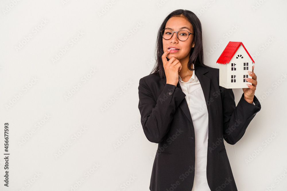 Young business latin woman holding a toy house isolated on white background relaxed thinking about something looking at a copy space. Young business latin woman holding a toy house isolated on white background relaxed thinking about something looking at a copy space.