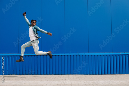 african guy jumping with a phone in his hands, in the city against the background of a blue building