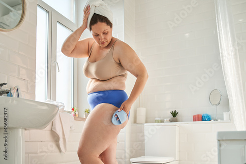 Woman with overweight body holding dry brush and massaging her hips attentively