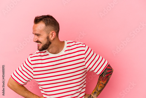 Young caucasian man with tattoos isolated on pink background laughs happily and has fun keeping hands on stomach.