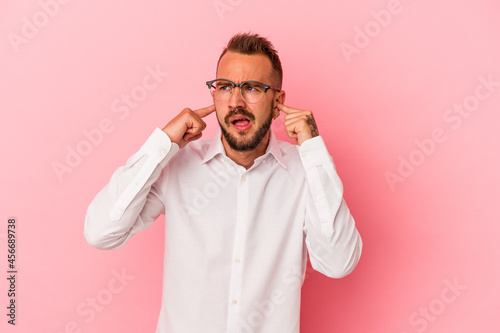 Fotografie Young caucasian man with tattoos isolated on pink background  covering ears with fingers, stressed and desperate by a loudly ambient