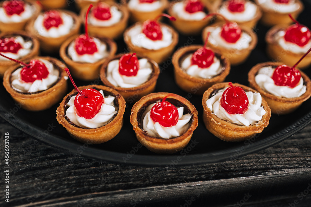 Close-up dough tartlets with cheese cream, cherries stand on a round tray, plate on a wooden table. Banquet, buffet, top view.