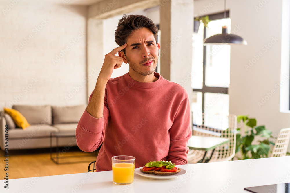 Young mixed race man having breakfast in his kitchen pointing temple with finger, thinking, focused on a task. Young mixed race man having breakfast in his kitchen pointing temple with finger, thinking, focused on a task.
