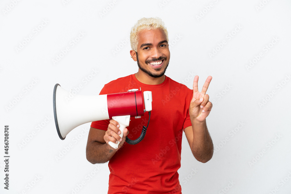 Young Colombian handsome man isolated on white background holding a megaphone and smiling and showing victory sign Young Colombian handsome man isolated on white background holding a megaphone and smiling and showing victory sign