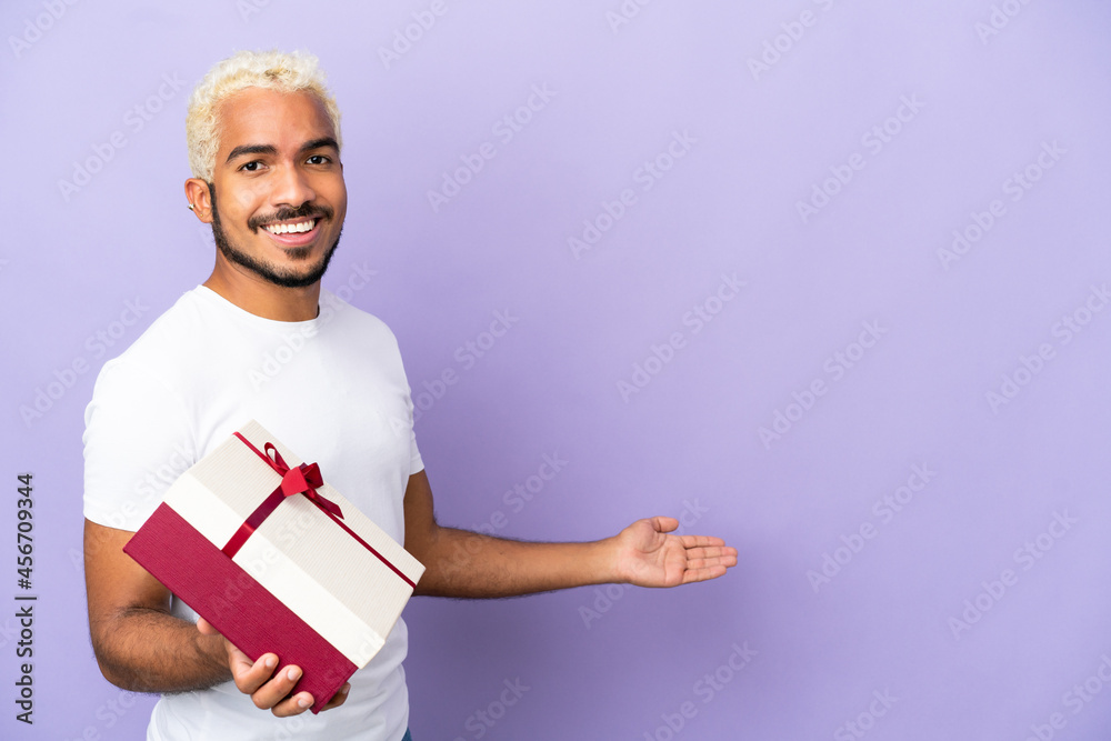 Young Colombian man holding a gift isolated on purple background extending hands to the side for inviting to come Young Colombian man holding a gift isolated on purple background extending hands to the side for inviting to come