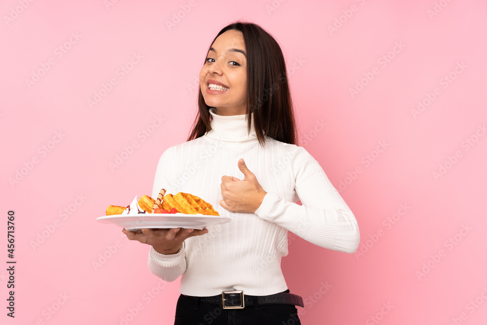 Young brunette woman holding waffles over isolated pink background giving a thumbs up gesture Young brunette woman holding waffles over isolated pink background giving a thumbs up gesture