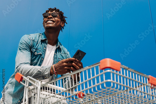 Afro black man with a shopping cart and a phone in his hands, makes purchases online, background blue