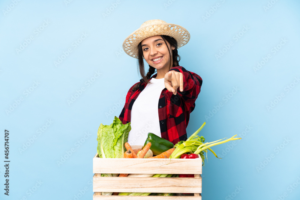 Young farmer Woman holding fresh vegetables in a wooden basket points finger at you with a confident expression