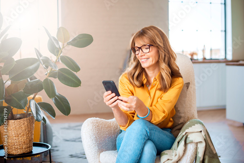Canvastavla Cheerful mature woman using mobile phone while relaxing in the armchair at home