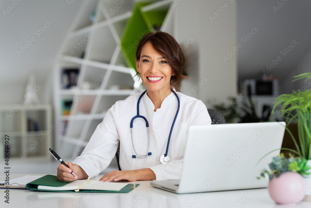 Smiling female doctor using laptop and writing something while sitting at doctor's office Smiling female doctor using laptop and writing something while sitting at doctor's office