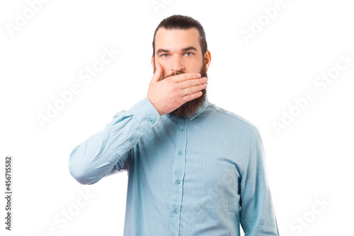 Serious young man is standing over white background while covering his mouth with his hand.