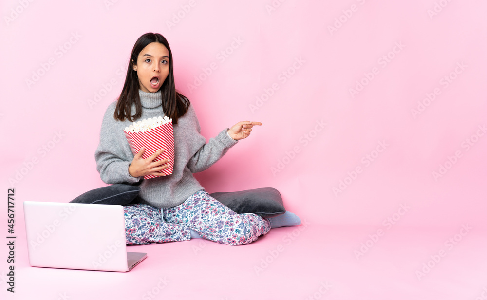 Young mixed race woman eating popcorn while watching a movie on the laptop with surprise expression while pointing side Young mixed race woman eating popcorn while watching a movie on the laptop with surprise expression while pointing side