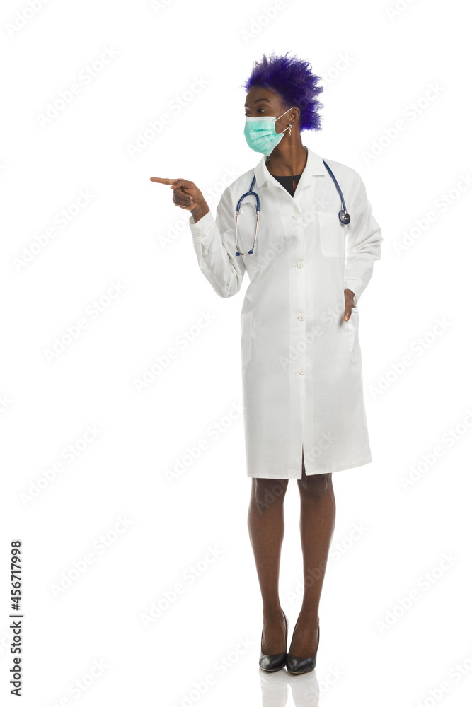 Black Female Doctor In Protective Face Mask Is Standing, Pointing To The Side And Looking Black Female Doctor In Protective Face Mask Is Standing, Pointing To The Side And Looking