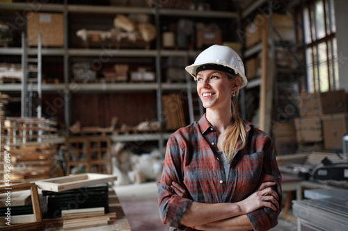 Portrait of mid-adult female carpenter standing in carpentery workshop, looking aside and smiling. Small business concept.