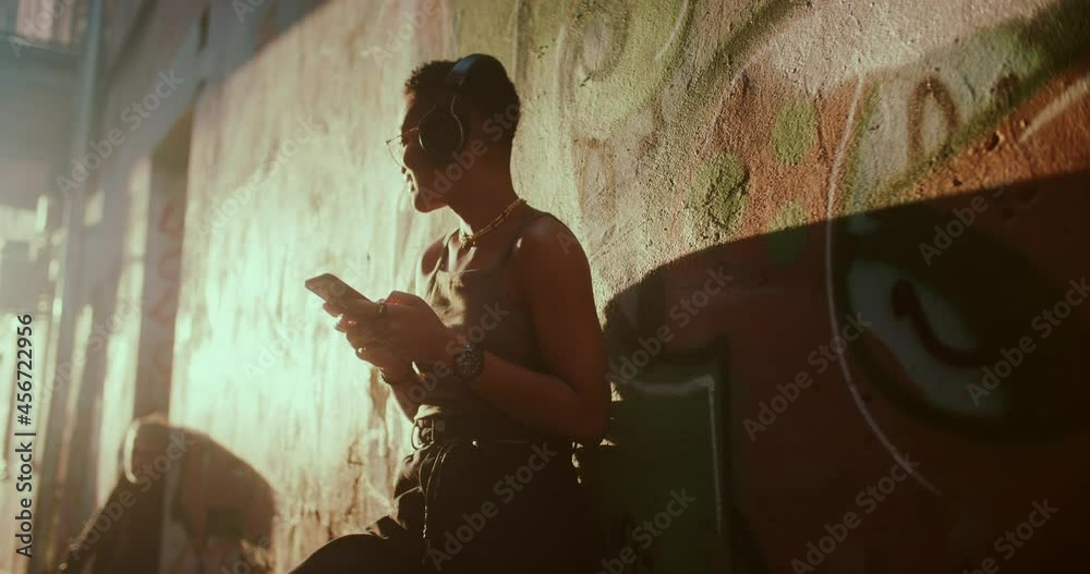 Joyful attractive young african girl in music headphones listening songs on mobile phone, enjoying dancing rhythm and sunny summer mood. Girl on street. Stylish people.