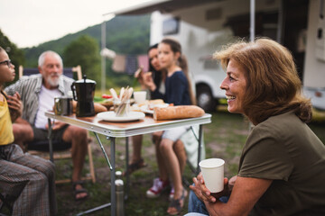 Multi-generation family sitting and eating outdoors by car, caravan holiday trip.