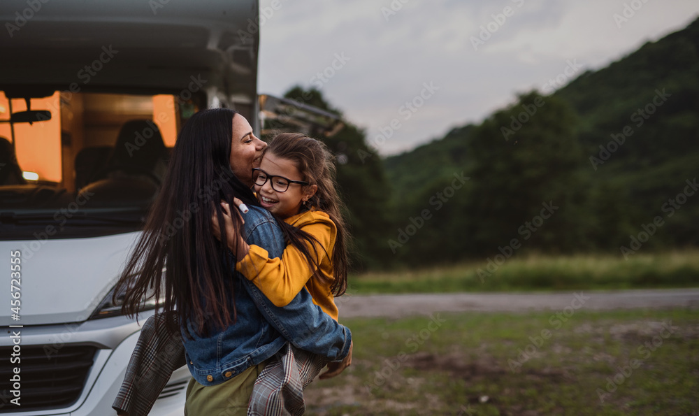 Happy mother with small daughter hugging by car outdoors in campsite at dusk, caravan family holiday trip. - obrazy, fototapety, plakaty 