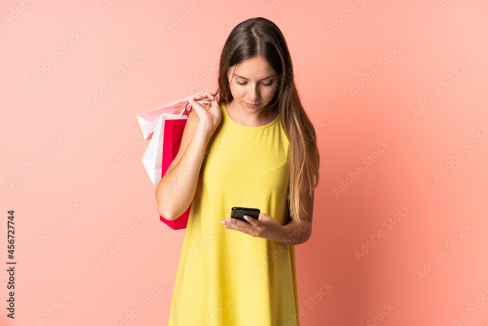 Young Lithuanian woman isolated on pink background holding shopping bags and writing a message with her cell phone to a friend Young Lithuanian woman isolated on pink background holding shopping bags and writing a message with her cell phone to a friend