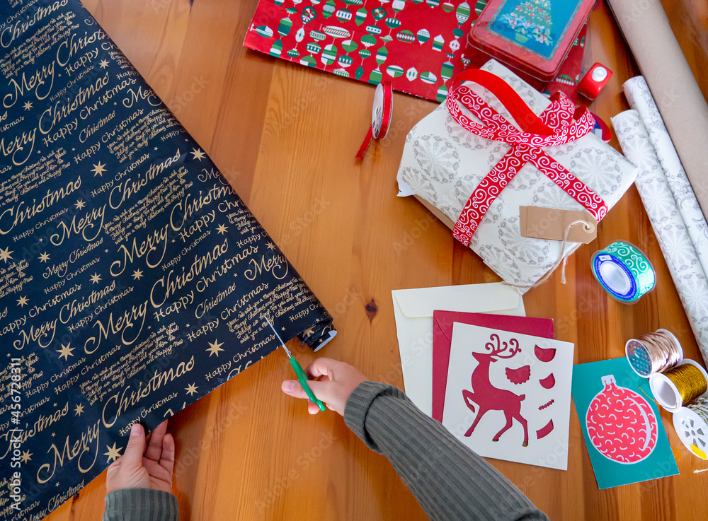 Fototapeta premium Woman hands cutting black Christmas wrapping paper. Work station for Xmas gifts decoration with ribbon, string and holiday cards. Top view of Christmas season flat lay with Christmas pattern