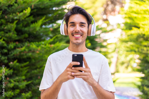 Young caucasian man in a park listening music with a mobile and looking front