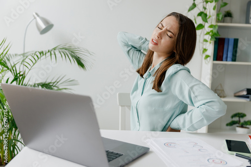 Young exhausted sick ill employee business woman in blue shirt hold hands on spine suffer from backache sit work at workplace desk with laptop pc computer at office indoors Achievement career concept photo