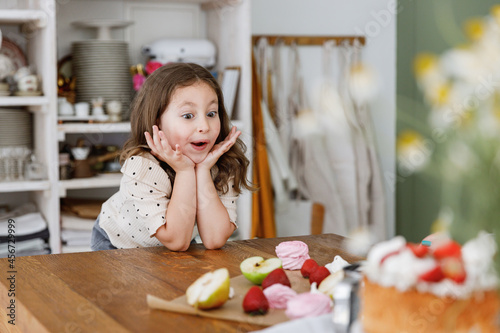 Young impressed amazed fun happy little kid child girl daughter sitting at table near fruits for sweet pie cake, learn how to cook in green kitchen indoors. People lifestyle, Mother's Day concept
