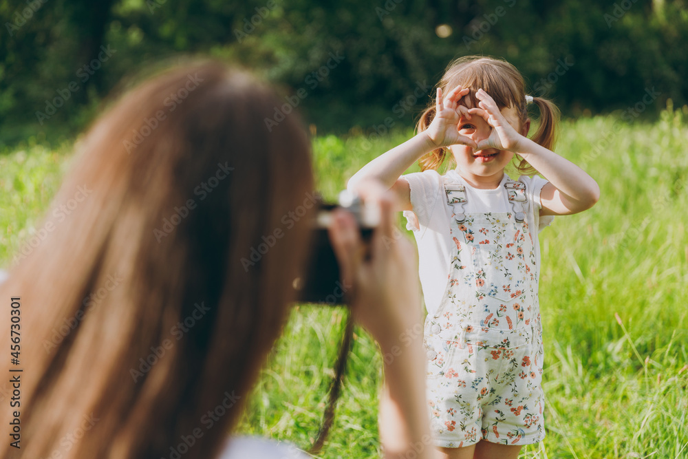 Obraz premium Photographer woman in white clothes take picture shot on photo camera of child baby girl 5-6 years old show heart-shape sign gesture Mommy rest with little kid daughter outdoor. Love family concept