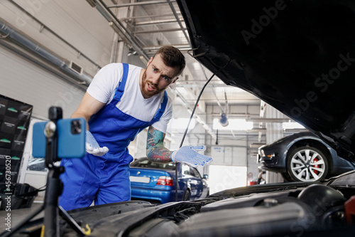 Young sad puzzled technician car mechanic man in blue overalls white t-shirt talk mobile cell on phone stand asking how to do fix problem with raised hood work in vehicle repair shop workshop indoors