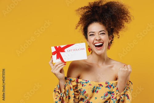 Young happy overjoyed excited fun woman 20s with culry hair in casual clothes hold gift certificate coupon voucher card for store do winner gesture isolated on plain yellow background studio portrait