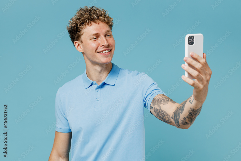 Smiling charismatic fun young curly man 20s years old wears azure t-shirt doing selfie shot on mobile phone post photo on social network isolated on plain pastel light blue background studio portrait. Smiling charismatic fun young curly man 20s years old wears azure t-shirt doing selfie shot on mobile phone post photo on social network isolated on plain pastel light blue background studio portrait.