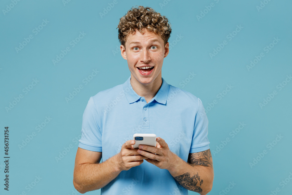 Surprised shocked vivid satisfied smiling overjoyed young curly man 20s years old wears azure t-shirt hold in hand use mobile cell phone isolated on plain pastel light blue background studio portrait Surprised shocked vivid satisfied smiling overjoyed young curly man 20s years old wears azure t-shirt hold in hand use mobile cell phone isolated on plain pastel light blue background studio portrait