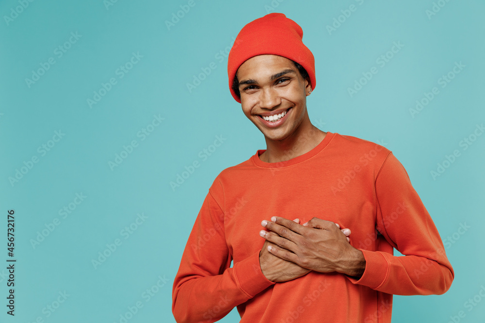 Young smiling happy thankful cheerful positive african american man in red shirt hat put folded hands on heart isolated on plain pastel light blue background studio portrait. People lifestyle concept. Young smiling happy thankful cheerful positive african american man in red shirt hat put folded hands on heart isolated on plain pastel light blue background studio portrait. People lifestyle concept.