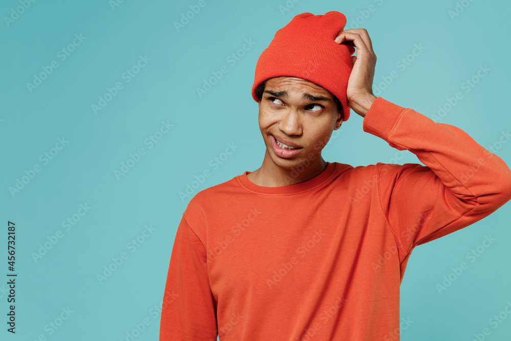 Young sad puzzled confused embarrassed african american man 20s in orange shirt hat look aside scratch head isolated on plain pastel light blue background studio portrait. People lifestyle concept