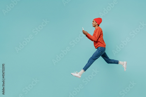Full body side view young happy african american man 20s in orange shirt hat jump high use mobile cell phone isolated on plain pastel light blue background studio portrait. People lifestyle concept.