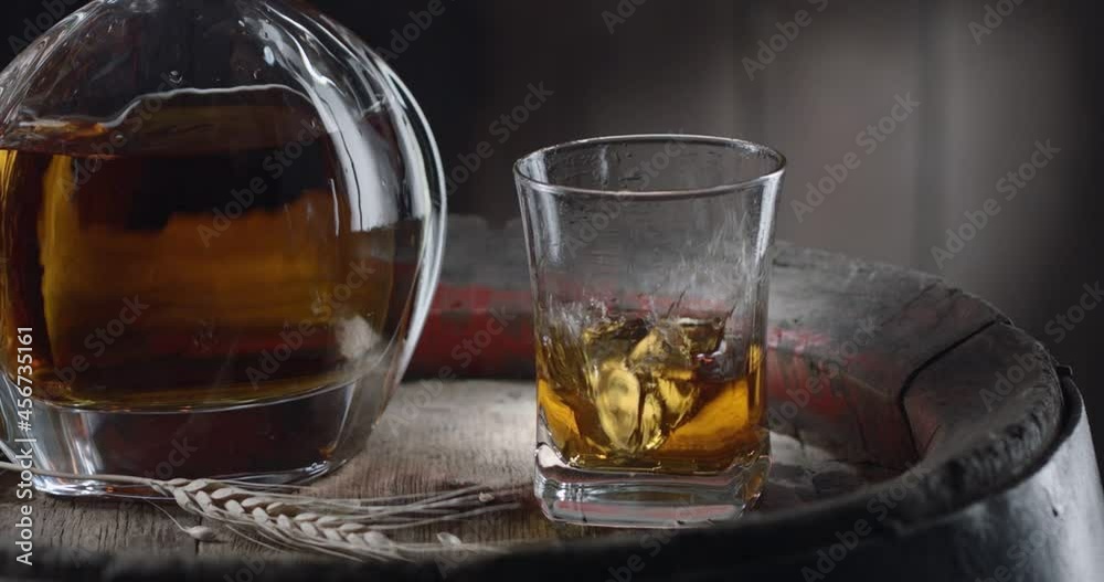 An ice cube slowly falls into a glass of whiskey on top of a vintage whiskey barrel. A decanter with a drink stands nearby, a dark brown background. Blackmagic Ursa Pro G2. An ice cube slowly falls into a glass of whiskey on top of a vintage whiskey barrel. A decanter with a drink stands nearby, a dark brown background. Blackmagic Ursa Pro G2.