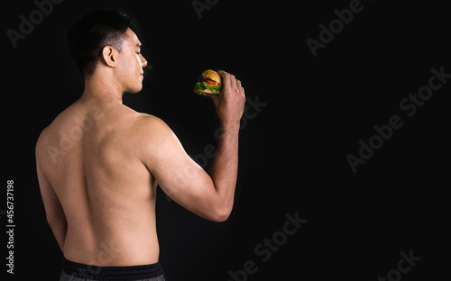 Asian sportive handsome strong man with shirtless, smiling with happiness, holding hamburger junk food and standing on black isolated background cutout. Health, Sport Concept.