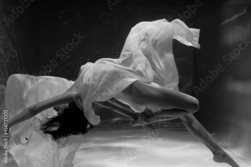 Black and white photo where a beautiful girl in a white dress swims underwater in the pool, and she looks like a siren or a mermaid