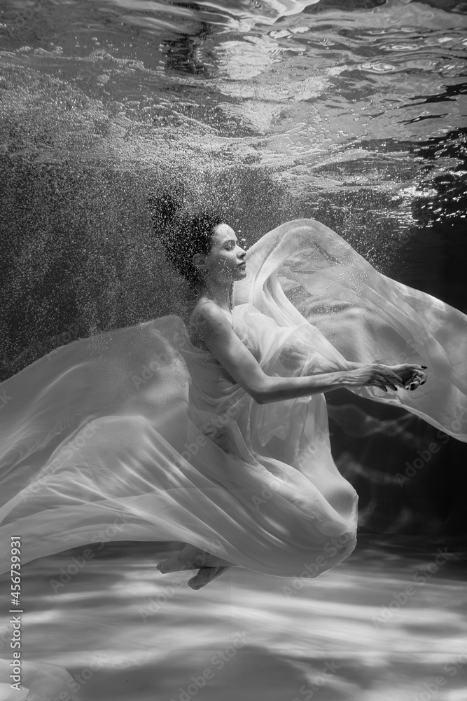 Black and white photo where a beautiful girl in a white dress swims underwater in the pool, and she looks like a siren or a mermaid Black and white photo where a beautiful girl in a white dress swims underwater in the pool, and she looks like a siren or a mermaid