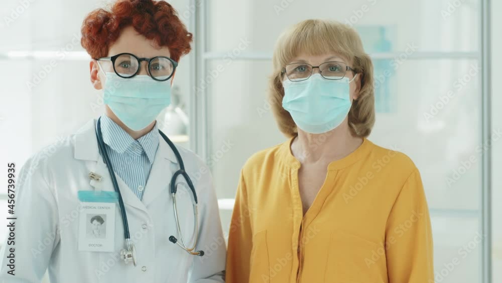 Portrait of young female doctor and elderly woman in protective masks standing together in clinic and posing for camera in time of coronavirus outbreak